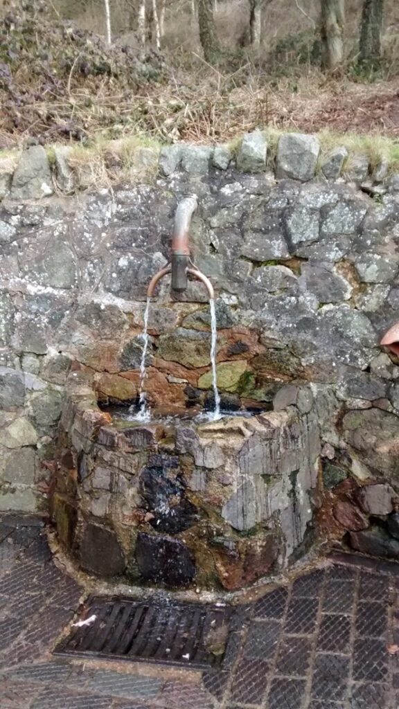 Photograph of water pouring from a waterspout onto rocks and to a drain, which provides water to the general public at a roadside location in central England.